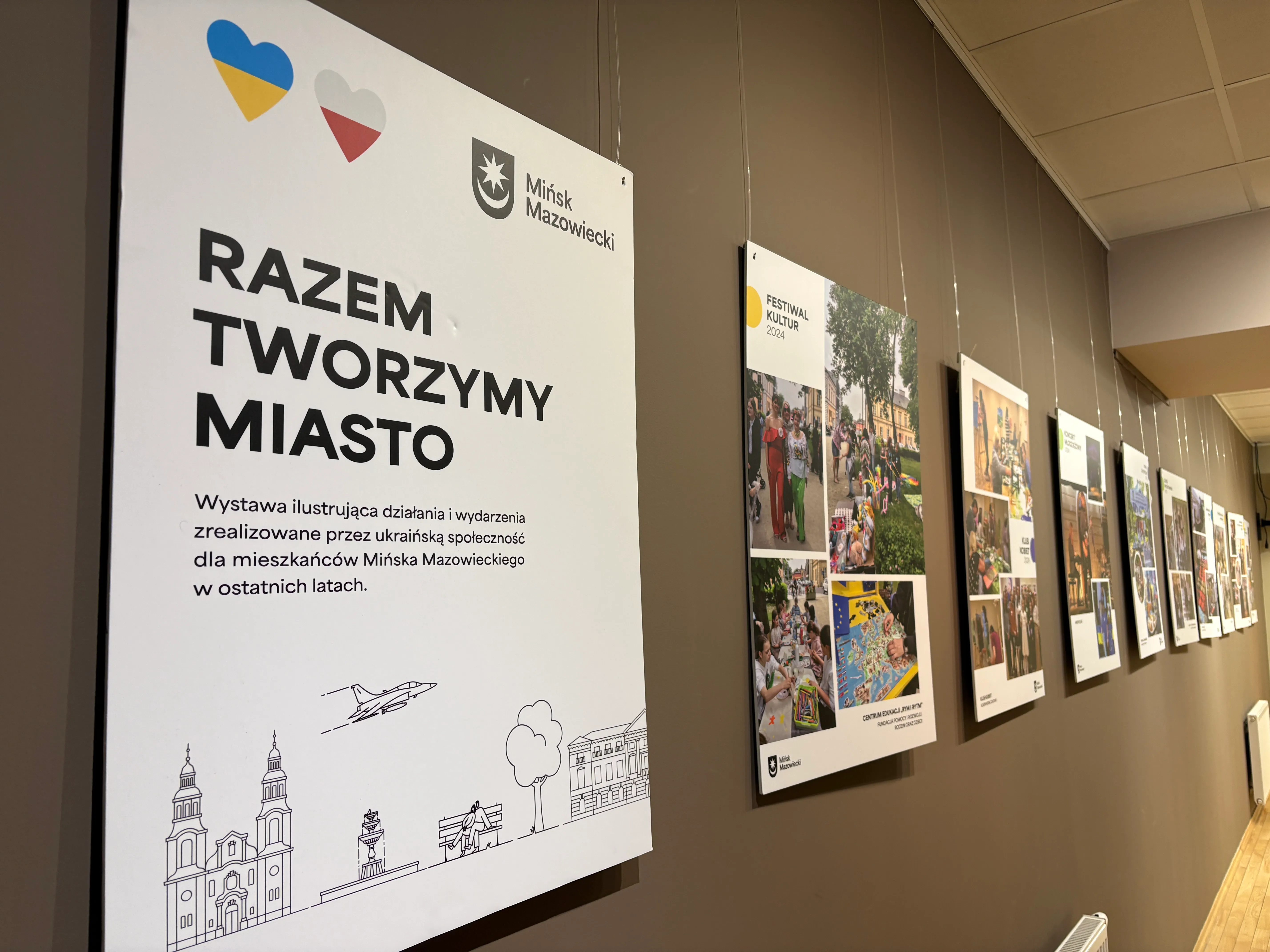 Kliknięcie powiększy obraz. Duży biały panel informacyjny z dwoma kolorowymi sercami w lewym górnym rogu zamocowany na brązowej ścianie. Po prawej stronie kilka wiszących plansz z kolażami fotografii ustawionych w rzędzie. Wnętrze galerii z podwieszanym sufitem i grzejnikiem przy podłodze.