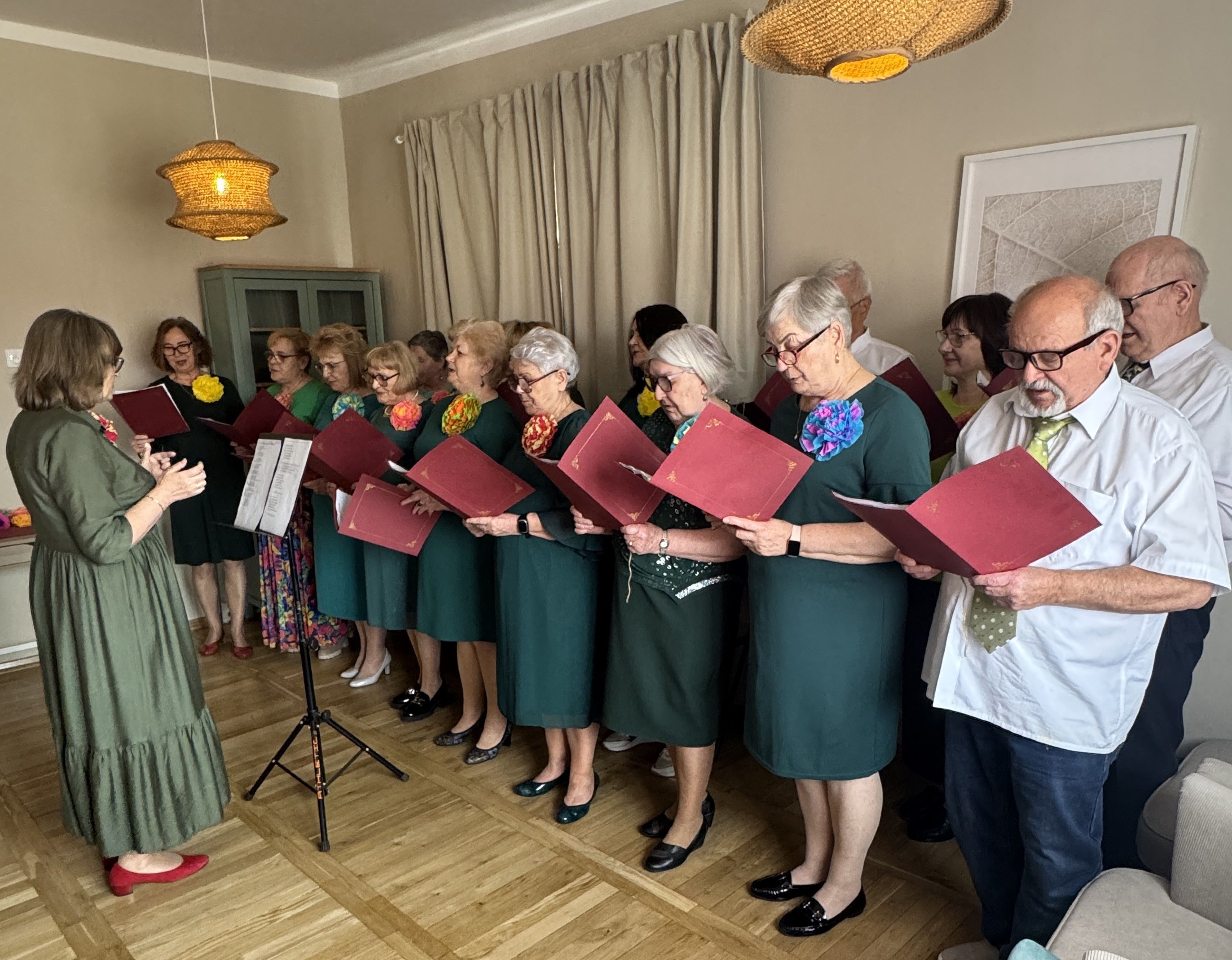 Kliknięcie powiększy obraz. Grupa około piętnastu starszych osób stoi w rzędzie w jasnym pokoju i śpiewa, trzymając czerwone teczki z nutami. Kobiety noszą zielone sukienki z kolorowymi kwiatami przy dekolcie po lewej, dyrygentka w zielonej sukni stoi z przodu po lewej. Dwaj mężczyźni w białych koszulach stoją po prawej, w tle jasne zasłony, szafka i plecione lampy sufitowe.
