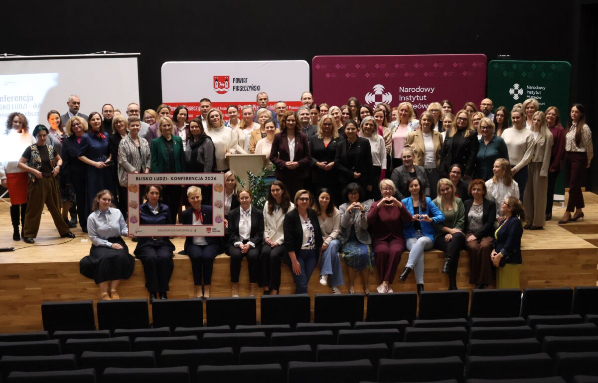 Kliknięcie powiększy obraz. Około sześćdziesięciu dorosłych osób stoi i siedzi na drewnianej scenie w sali konferencyjnej. Większość to kobiety, jest kilka mężczyzn; w tle wiszą białe, bordowe i zielone banery z logotypami.