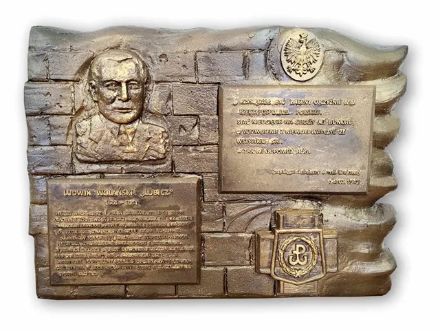 Kliknięcie powiększy obraz. Miedziana pamiątkowa płaskorzeźba na tle stylizowanej ceglanej ściany. W górnym lewym rogu znajduje się reliefowe popiersie mężczyzny. Po prawej u góry umieszczony jest okrągły orzeł państwowy. Poniżej orła i po lewej widoczne są dwie prostokątne tablice z inskrypcjami. W prawym dolnym rogu znajduje się tarcza z symbolem Kotwicy i falującą dekoracyjną wstęgą. Całość ma metaliczny brązowy kolor.