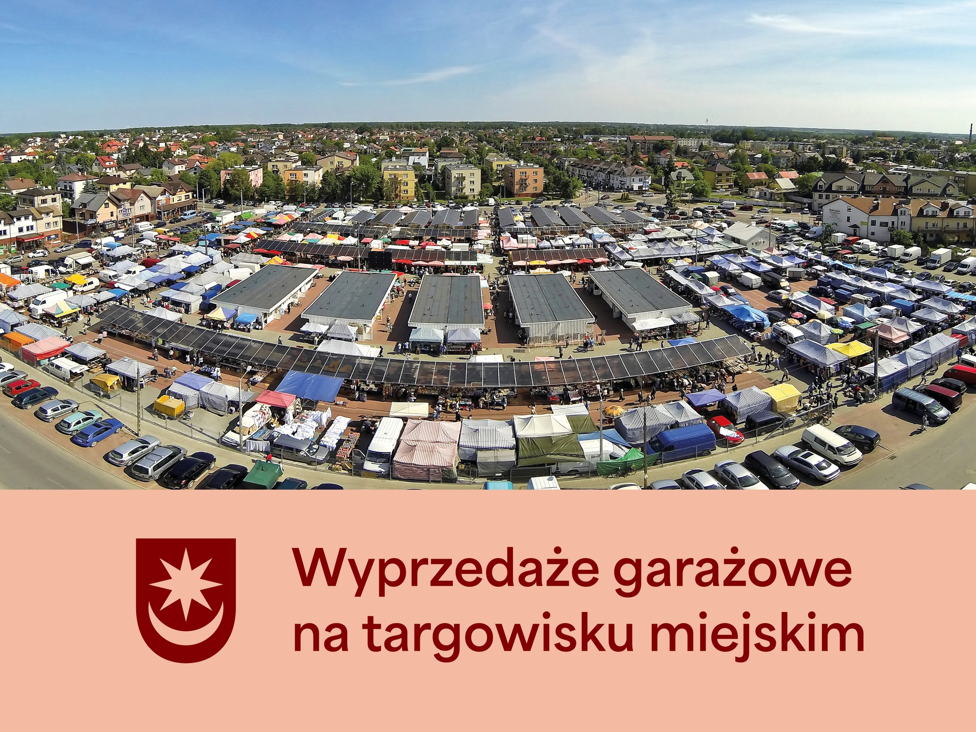 Kliknięcie powiększy obraz. Targ miejski z wieloma namiotami i stoiskami ustawionymi w rzędach na otwartym parkingu. W tle niskie budynki mieszkalne i drzewa pod jasnym, niebieskim niebem w ciągu dnia.