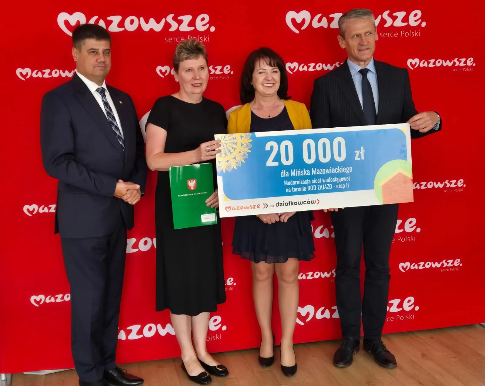 Kliknięcie powiększy obraz. Cztery osoby stoją razem na tle czerwonego baneru z napisem "Mazowsze, serce Polski", trzymając duży czek na kwotę 20 000 zł oraz dokumenty związane z projektem modernizacji sieci wodociągowej.