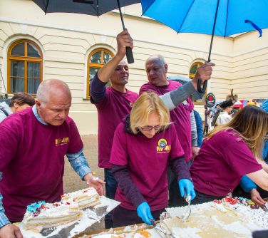 Na dworze, dzień. Pod parasolkami przed palacem osoby w bordowych koszulkach kroją tort