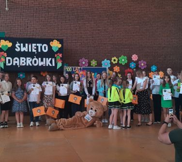 grupa osób pozuje do zdjęcie, przed nimi leży duża maskotka niedźwiedzia, w tle na ścianie kolorowe kwaiaty i napis święto...