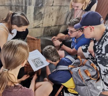 grupa mlodych ludzi pochyla się nad wiklinową skrzynią, na której leżą kartki z tekstem i zdjciem drzwi