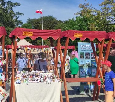 Dwór. Drewniane stragany z czerwonymi daszkami z napisem muzeum ziemi mińskiej. W tle drzewa i maszt z flagą biało-czerwoną