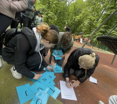 Na dworze. Grupa osób kuca na trawie. pochylają się nad niebieskimi kartkami