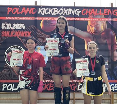 Pomieszczenie. Trzy nastolatki na podium z dyplomami w rękach i medalami na szyi. W tle na drabinkach duży baner