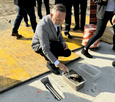 Na dworze, słonecznie. Mężczyzna kuca. Kielnią nabiera cement z plastikowego pojemnika