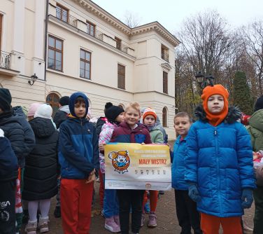 Na dworze, dzień. Przed pałacem grupa dzieci w kurtkach i czapkach. Chłopiec trzyma baner Mały mistrz