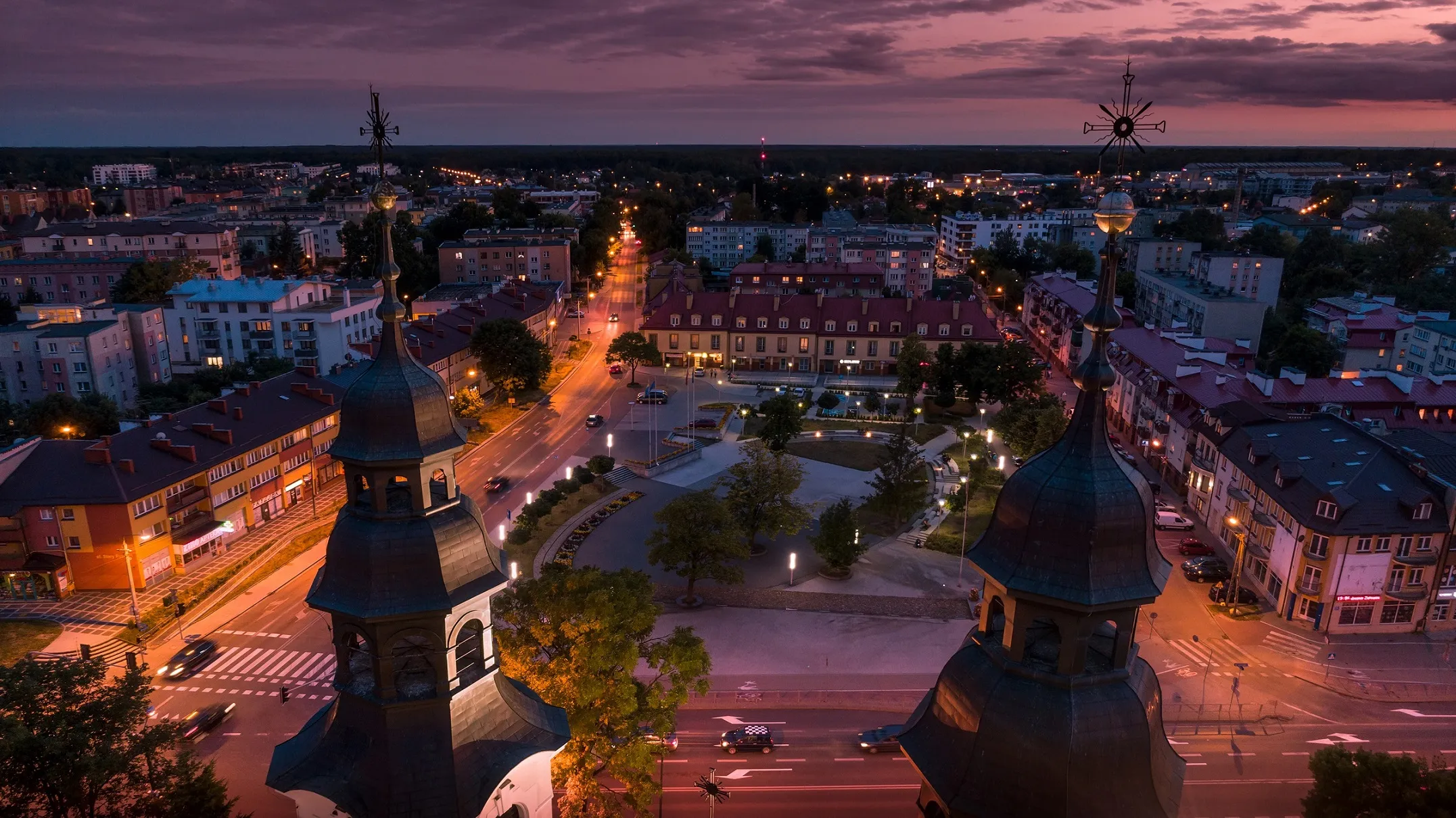 Dwa wieże kościoła z krzyżami na szczytach na pierwszym planie oraz oświetlona ulica i plac miejski z budynkami w tle podczas zmierzchu.