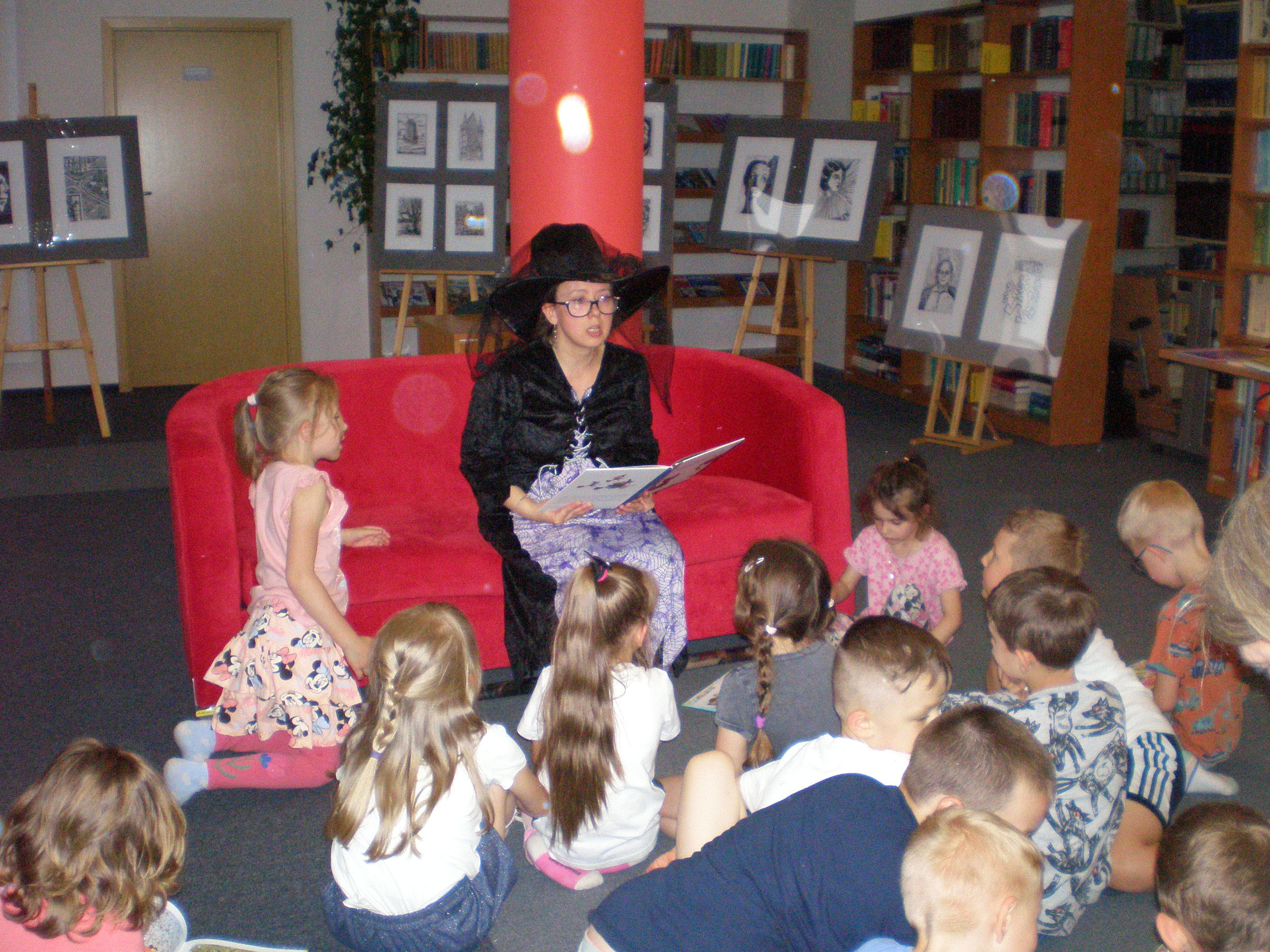 Sala biblioteczna. Na czerwonej sofie siedzi kobieta w czarnym kapeluszu. Czyta zgromadzonym przed nią dzieciom